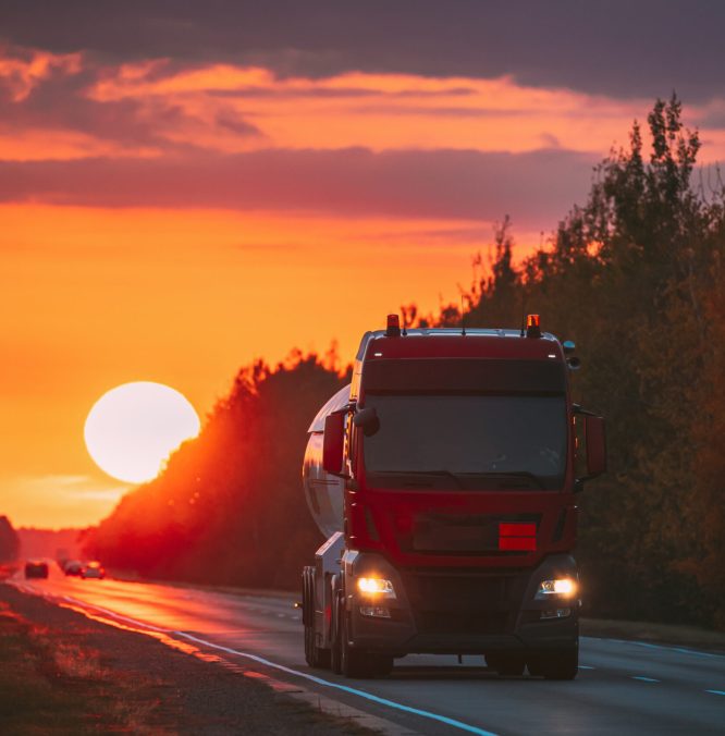 Red Truck Or Tractor Unit, Prime Mover, Traction Unit In Motion On Road, Freeway. Asphalt Motorway Highway Against Background Of Big Sunset Sun. Business Transportation And Trucking Industry